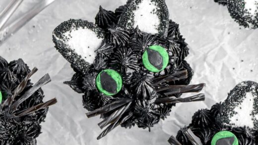 overhead image of black cat cupcake with seven more cupcakes around, two forks, and white flowers beside.