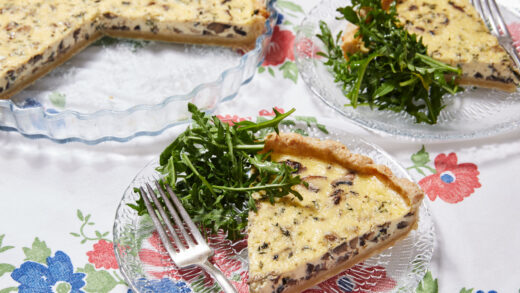 Two slices of Mushroom and Thyme Leaf Tart are cut from the whole tart and served on clear glass plates with forks. The tart has golden, buttery and crumbly crust loaded with velvety custard filling and mushrooms, paired with green earthy thyme leaves. The rest of the tart is in the tart dish.