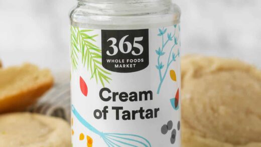 A jar of cream of tartar on a countertop next to a teaspoonful, with sugar cookies in the background.