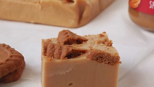 Close up of a piece of Biscoff Fudge on a sheet of baking paper. In the background is more fudge and jar of Biscoff spread.