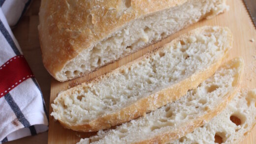 Overnight crusty bread partially sliced,  placed on a wooden board with a knife & kitchen towel in the background.