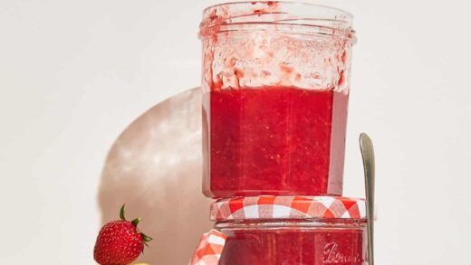 two jars of strawberry jam stacked with a lemon and strawberry beside them