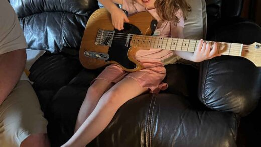 Little girl sitting on a brown leather couch holding a guitar.