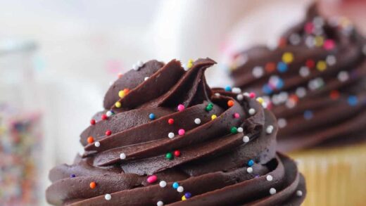 Close up of a yellow cupcake decorated with chocolate frosting and sprinkles in an open cupcake wrapper, with more cupcakes in the background.