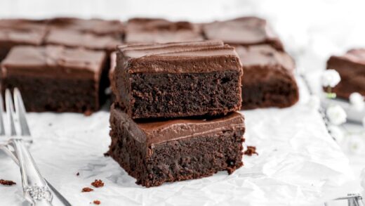 two stacked frosted brownies on parchment lined sheet pan with two forks beside and more brownies behind.