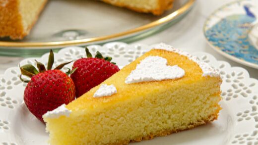 A slice of the Orange Almond Tea Cake sits on a white plate with 2 strawberries and the rest of the cut cake is behind it.