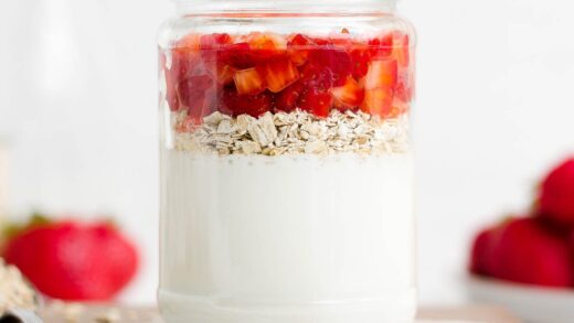 a glass jar holding the layered ingredients used to make healthy strawberry protein overnight oats, while resting on a wooden cutting board alongside a spoonful of oats – easy overnight oatmeal recipe – high protein, gluten free, sugar free, low fat, clean eating – made with Greek yogurt, oats, fresh strawberries, and no added sugar