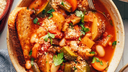 Bowls of veggie white bean minestrone topped with vegan parmesan cheese and parsley and served with toasted bread